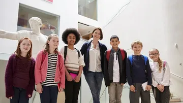 Ein Erwachsener und vier Kinder sitzen an einem Tisch und arbeiten an einem Projekt. Sie tragen bunte Kleidung. Der Raum ist hell mit Fenstern. Auf dem Tisch liegen Notizbücher.