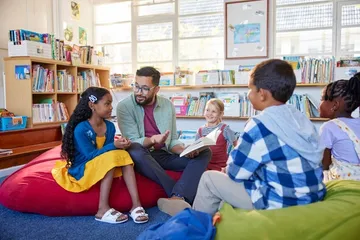 Ein Mann liest Kindern in einer Bücherei vor. Alle sitzen gemeinsam auf Sitzkissen und hören gespannt zu.