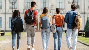 Fünf junge Leute gehen über einen Weg vor einem großen Gebäude. Sie tragen Rucksäcke und lässige Kleidung wie Jeans und T-Shirts. Das Gebäude hat viele Fenster und sieht alt aus. Die Sonne macht die Szene hell und freundlich.