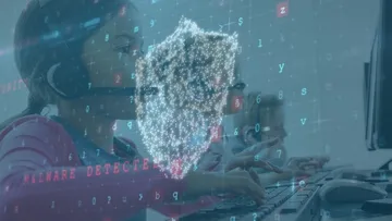 Eine Person tippt am Computer, über dem ein digitales Sicherheitssymbol erscheint.
