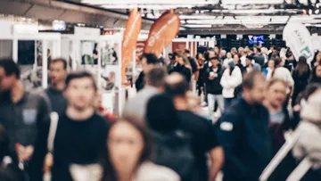 Ein geschäftiges Bild einer Menschenmenge auf einer Messe. Die Leute sind unscharf und tragen dunkle Kleidung. Über ihnen hängen orangefarbene Fahnen mit Logos. Im Hintergrund sieht man Glaswände und Bildschirme.