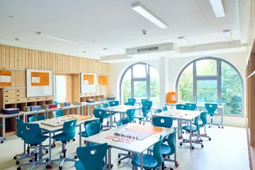 Heller und moderner Klassenraum mit großen, halbrunden Fenstern, die viel Tageslicht hereinlassen und den Blick ins Grüne freigeben. Mehrere Gruppentische mit blauen Stühlen auf Rollen sind im Raum verteilt. An der Rückwand befinden sich Holzverkleidungen mit Schubladen und weißen Tafeln, an denen orangefarbene Plakate hängen. Der Raum wirkt freundlich, aufgeräumt und flexibel für unterschiedliche Lernformen.