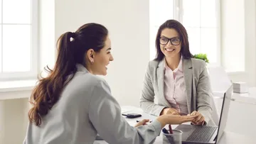 Zwei Frauen reden lächelnd am Schreibtisch miteinander.