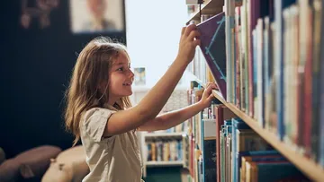 Kinder bauen an einem Tisch mit einem Laptop und Elektronikteilen. Eines hält ein Modellauto hoch. Sie tragen bunte Kleidung. Der Raum ist hell und mit Regalen ausgestattet.