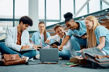 Studenten in blauen Kitteln sitzen auf dem Boden und lernen zusammen mit Laptop und Notizbüchern. Sie lächeln und zeigen aufeinander, Rucksäcke und Bücher liegen herum. Der Raum ist hell mit Fenstern.