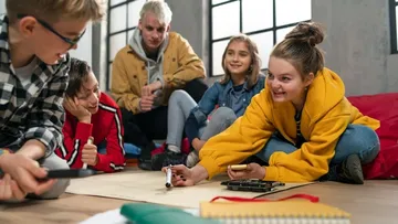 Drei Personen sammeln Müll in einem Park mit Bäumen. Ein Kind mit pinker Mütze füllt einen roten Sack. Eine Erwachsene und ein weiteres Kind helfen mit.