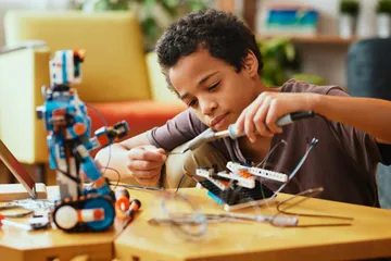 Ein Kind baut einen Roboter mit Bausteinen und Drähten. Dafür nutzt es eine Pinzette. Weitere Werkzeuge und ein Laptop liegen auf dem Tisch. Der Raum ist hell mit gelben Möbeln und Pflanzen.